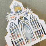 3d pop up greeting card of a white wedding church pattern with a happily ever after banner displayed on a wooden table